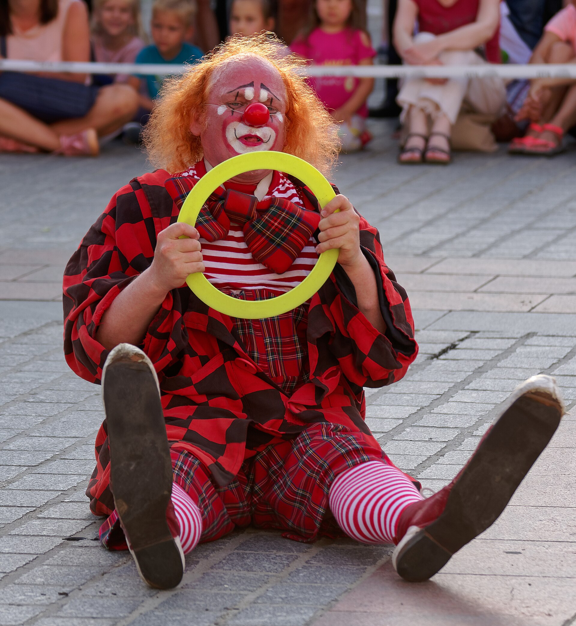 A clown sits on a paved surface holding a yellow plastic ring during a street performance.